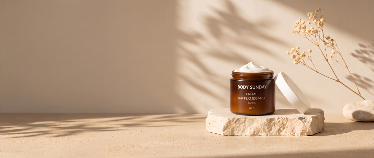 Pot ouvert de crème raffermissante corps Body Sunday posé sur une pierre claire, fond beige et fleurs séchées
